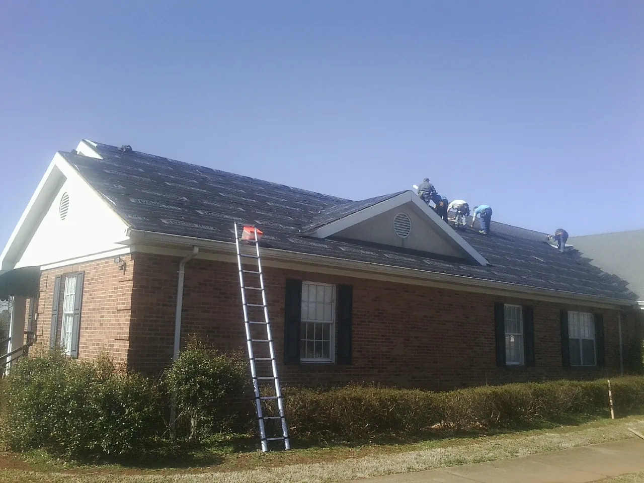 Crew tearing shingles off a brick ranch roof with a ladder set — Emergency Roof Repair work by our crew in Red Rock