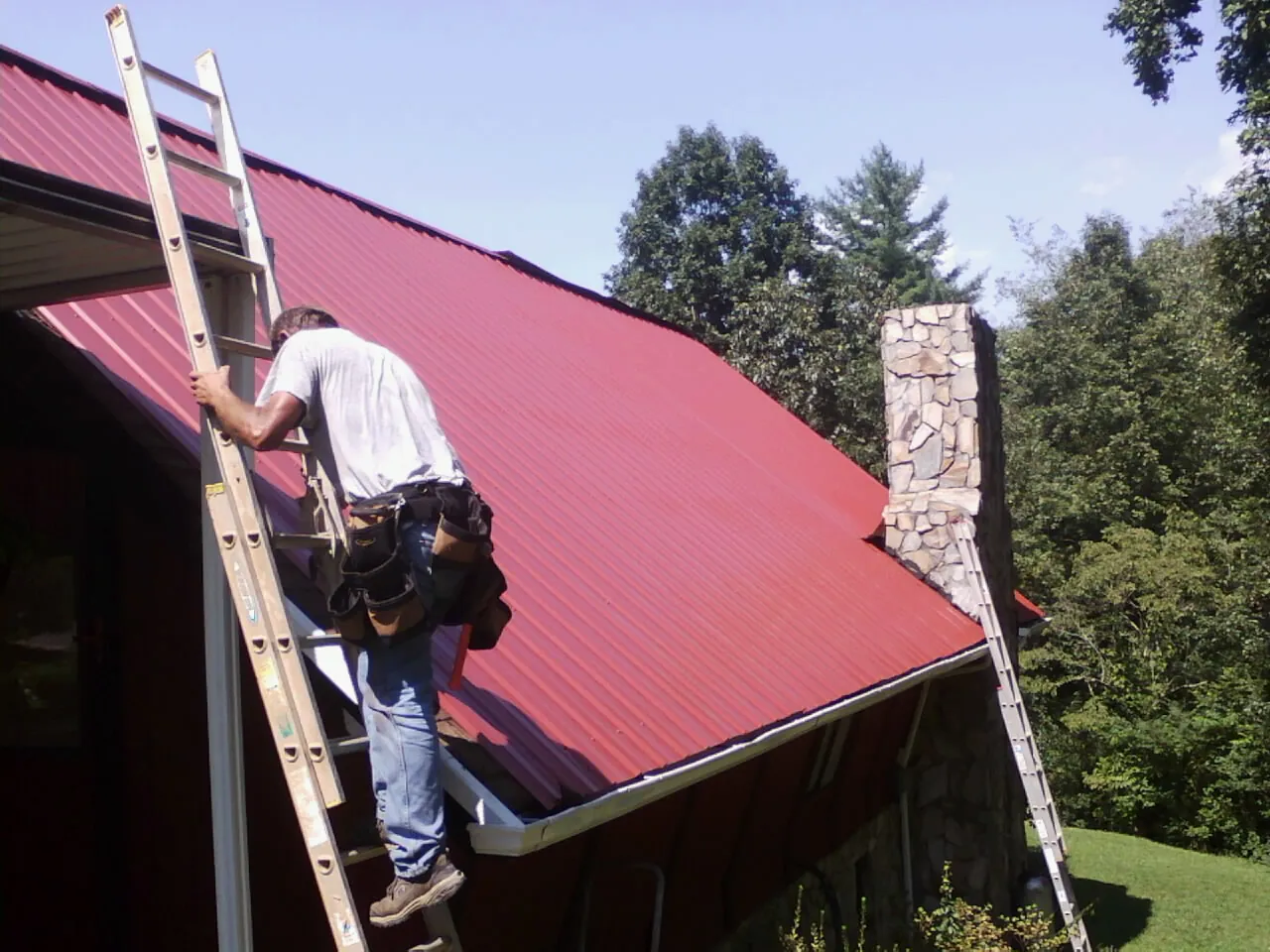 Roofer on a ladder beside a red metal roof and stone chimney — Soffit & Fascia Repair by Roofing Brothers in Red Rock
